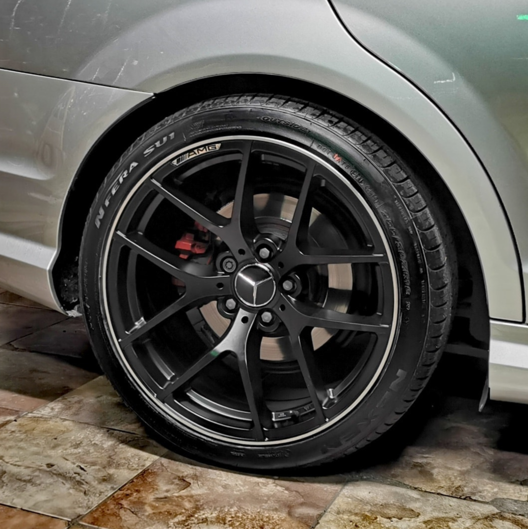 A black, multi-spoke car wheel with a Mercedes logo is stationary, set against a clean, tiled floor and silver vehicle body. Tire text reads: "N'FERA SU1," "AMG."