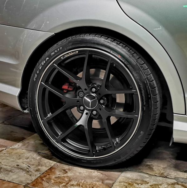 A black, multi-spoke car wheel with a Mercedes logo is stationary, set against a clean, tiled floor and silver vehicle body. Tire text reads: "N'FERA SU1," "AMG."