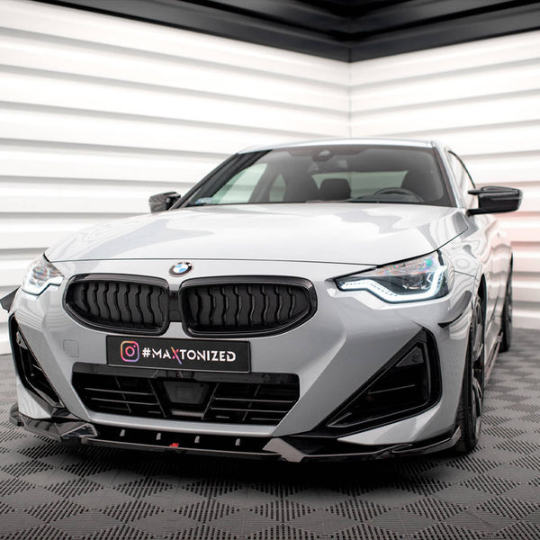 Car parked in a modern showroom with geometric wall designs, featuring a gray BMW with a sleek front grille and illuminated headlights. The license plate reads "#MAXTONIZED."