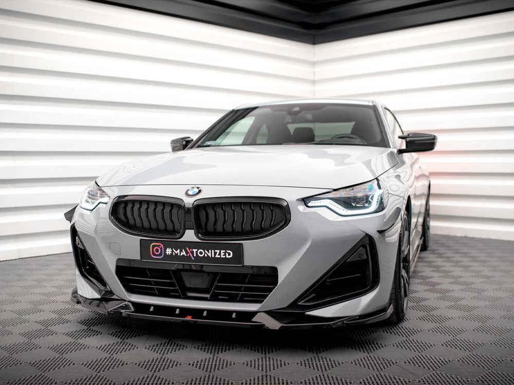 Car parked in a modern showroom with geometric wall designs, featuring a gray BMW with a sleek front grille and illuminated headlights. The license plate reads "#MAXTONIZED."
