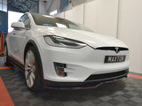 A white car with a Tesla logo is parked indoors on a checkered floor, displaying the license plate 