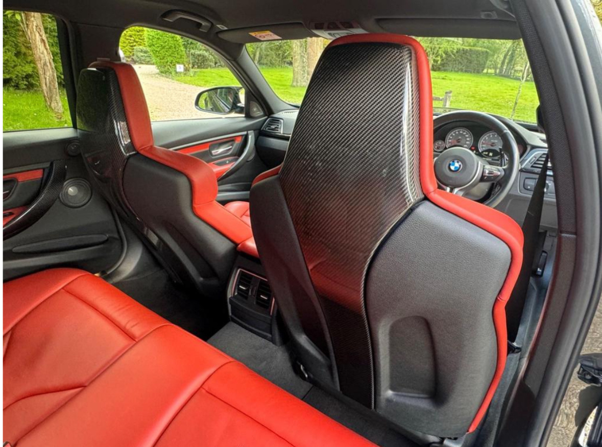 Car interior features red leather seats with carbon fiber backrests, viewed from the back seat; a steering wheel with a BMW logo is visible through the windshield.
