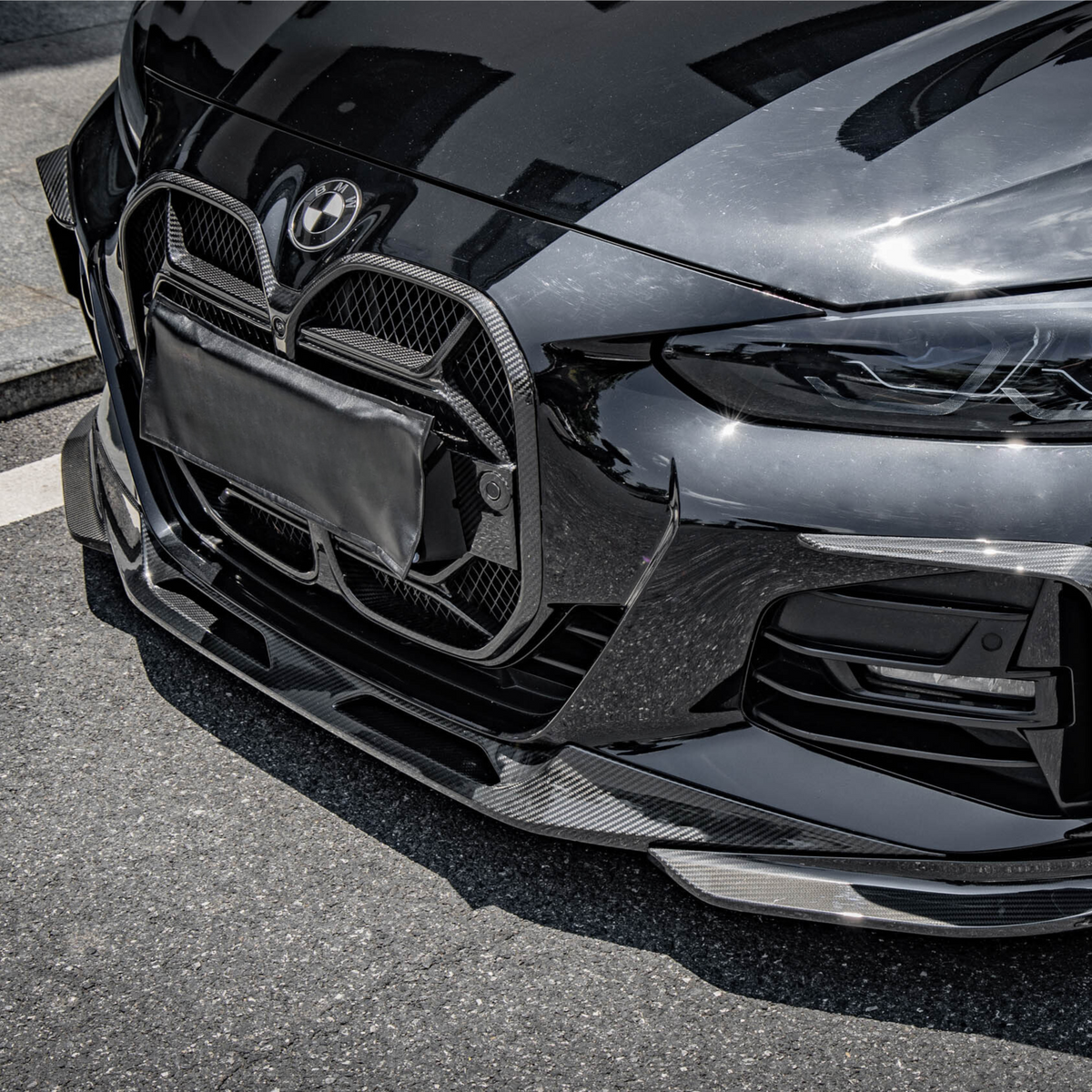 A front view of a sleek black car with a prominent grille. The vehicle is parked on a paved street, reflecting light and nearby structures.