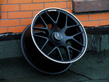 A black alloy wheel with a silver edge rests against an orange brick wall on a dark surface. The central cap features a three-pointed star logo and 