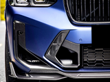 The close-up of a blue car front highlights a sleek headlight and sporty grille with carbon fiber detailing, parked on a road with a blurred background.
