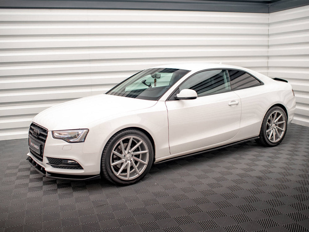 A white Audi coupe is parked, showcasing its sleek design with silver alloy wheels, in a well-lit room with textured gray flooring and horizontal white paneling on the walls.