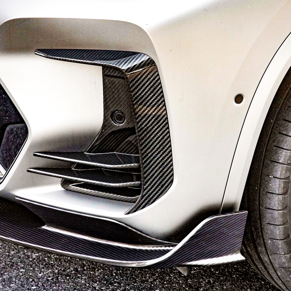 A car front bumper with carbon fiber accents and an alloy wheel is situated on an asphalt surface, showcasing aerodynamic features and a sporty design.