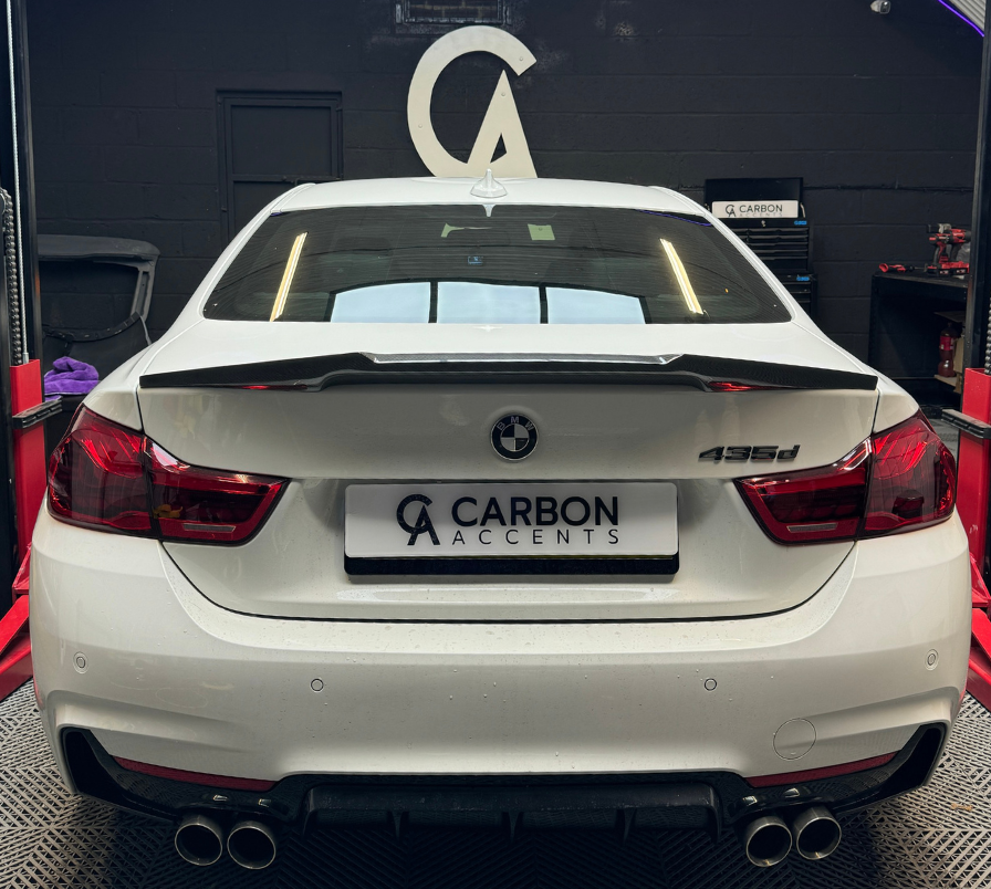 A white BMW 435d with a custom spoiler is parked on a vehicle lift inside an automotive workshop. The license plate reads "CARBON ACCENTS" with a logo above.