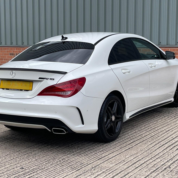 A white Mercedes-Benz AMG car is parked on a textured concrete surface beside a brick and corrugated metal wall. The car's license plate text is unclear. Black wheels and a rear spoiler enhance its sporty look.