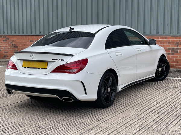 A white Mercedes-Benz AMG car is parked on a textured concrete surface beside a brick and corrugated metal wall. The car's license plate text is unclear. Black wheels and a rear spoiler enhance its sporty look.