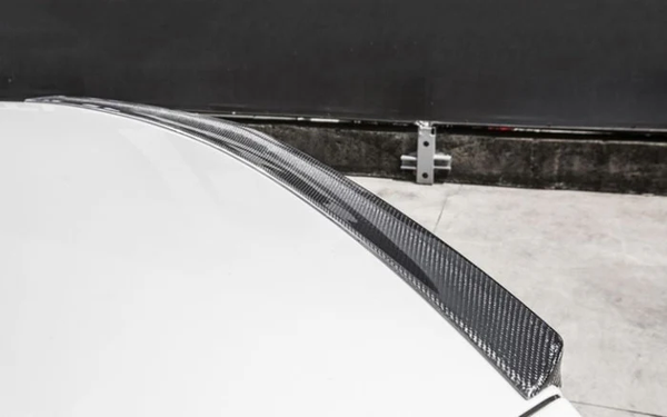 A carbon fiber car spoiler on a white vehicle is shown close-up, situated in an outdoor setting near a concrete wall and metal barrier post.