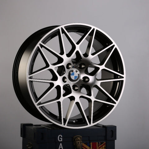 A silver and black alloy wheel with intricate geometric spokes rests on a blue crate with partial text "GA" and a visible UK flag. BMW logo is at the center.
