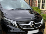 A black Mercedes-Benz van is parked on a gravel driveway, with a traditional brick house in the background. The front grille displays the brand's emblem prominently.