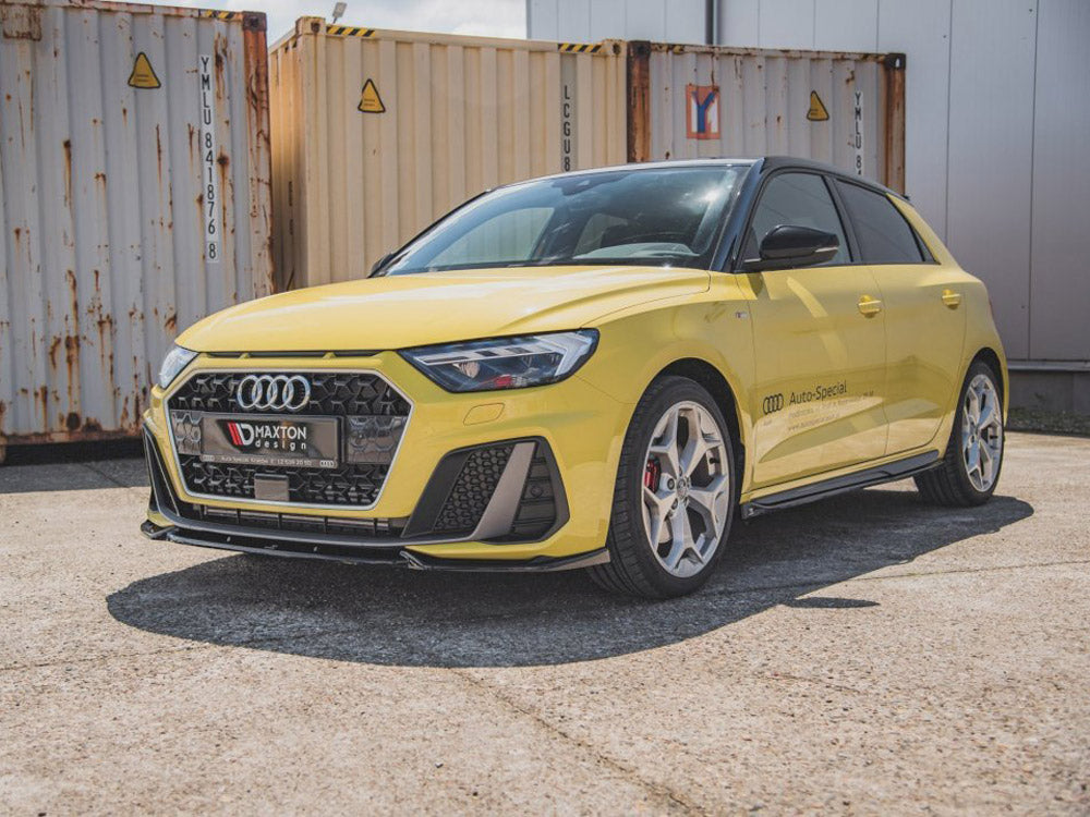 A yellow Audi car parked on concrete, with black trim and a hexagonal grille, displays "Maxton design" on the front. "AUDI Auto-Special" is inscribed on the side, next to shipping containers.