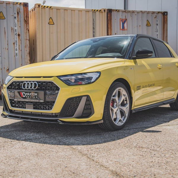 A yellow Audi car parked on concrete, with black trim and a hexagonal grille, displays "Maxton design" on the front. "AUDI Auto-Special" is inscribed on the side, next to shipping containers.