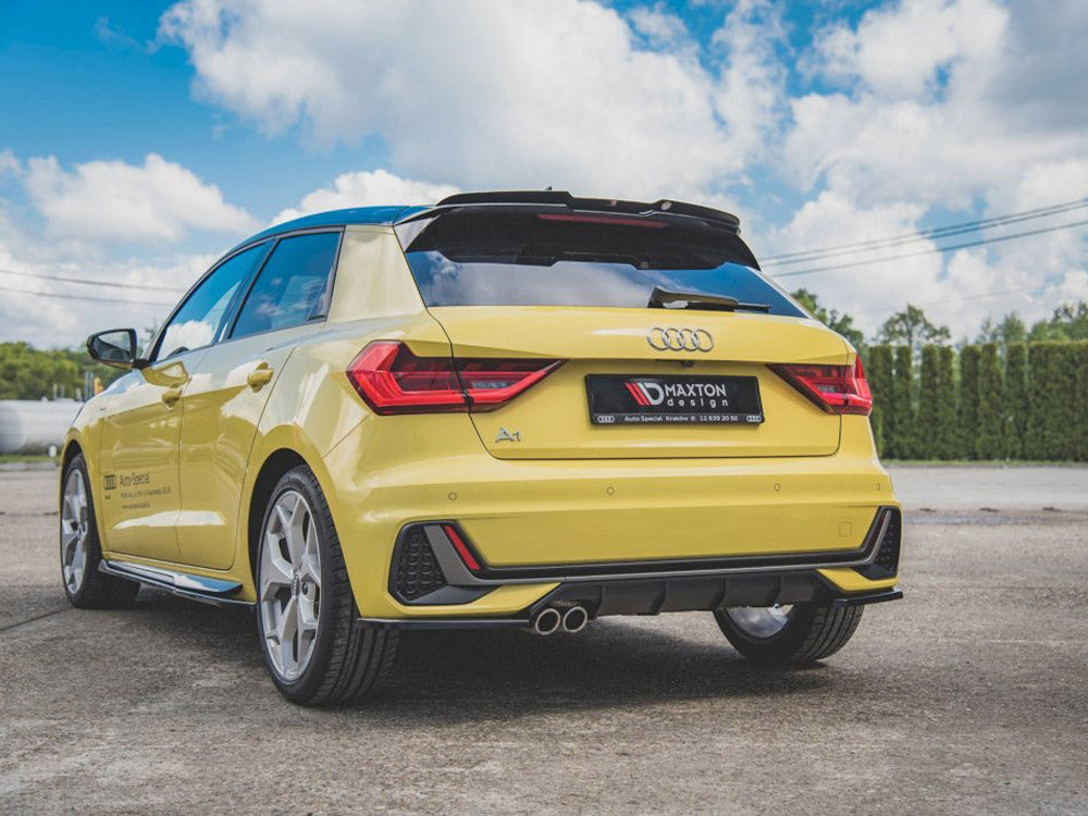 A yellow Audi A1 is parked on a concrete surface, featuring red tail lights and dual exhaust pipes. It has a license plate reading "MAXTON design." The sky is partly cloudy.