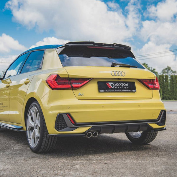 A yellow Audi A1 is parked on a concrete surface, featuring red tail lights and dual exhaust pipes. It has a license plate reading "MAXTON design." The sky is partly cloudy.