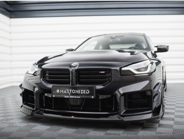 A black sports car with a sleek, aerodynamic design is parked on a textured floor in a well-lit garage. The license plate reads "#MAXTONIZED Dealer BMW M//Cars."