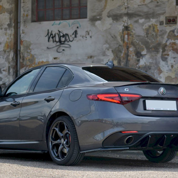 A sleek gray car is parked on a paved surface, its rear facing a weathered, graffiti-marked wall with peeling paint.