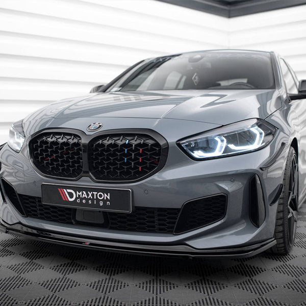 A sleek, grey BMW car with illuminated headlights sits on a checkered floor in a modern, white-panelled room. The license plate reads "MAXTON design."
