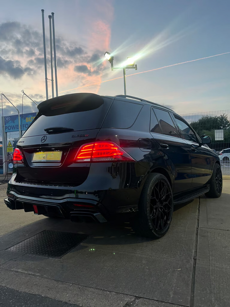A black Mercedes-Benz GL 350 SUV is parked on a concrete surface at dusk, with its rear lights glowing and streetlights illuminating the area against a cloudy sky.