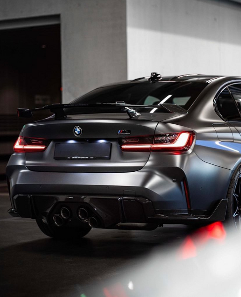 A sleek BMW car is parked in a dimly lit garage with a matte finish, featuring illuminated taillights and a prominent spoiler. The area appears modern and industrial.