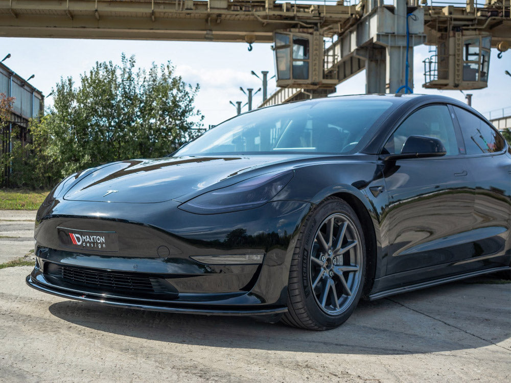 A sleek black car is parked on a concrete surface near industrial structures and greenery. The front bumper displays a "Maxton Design" logo.