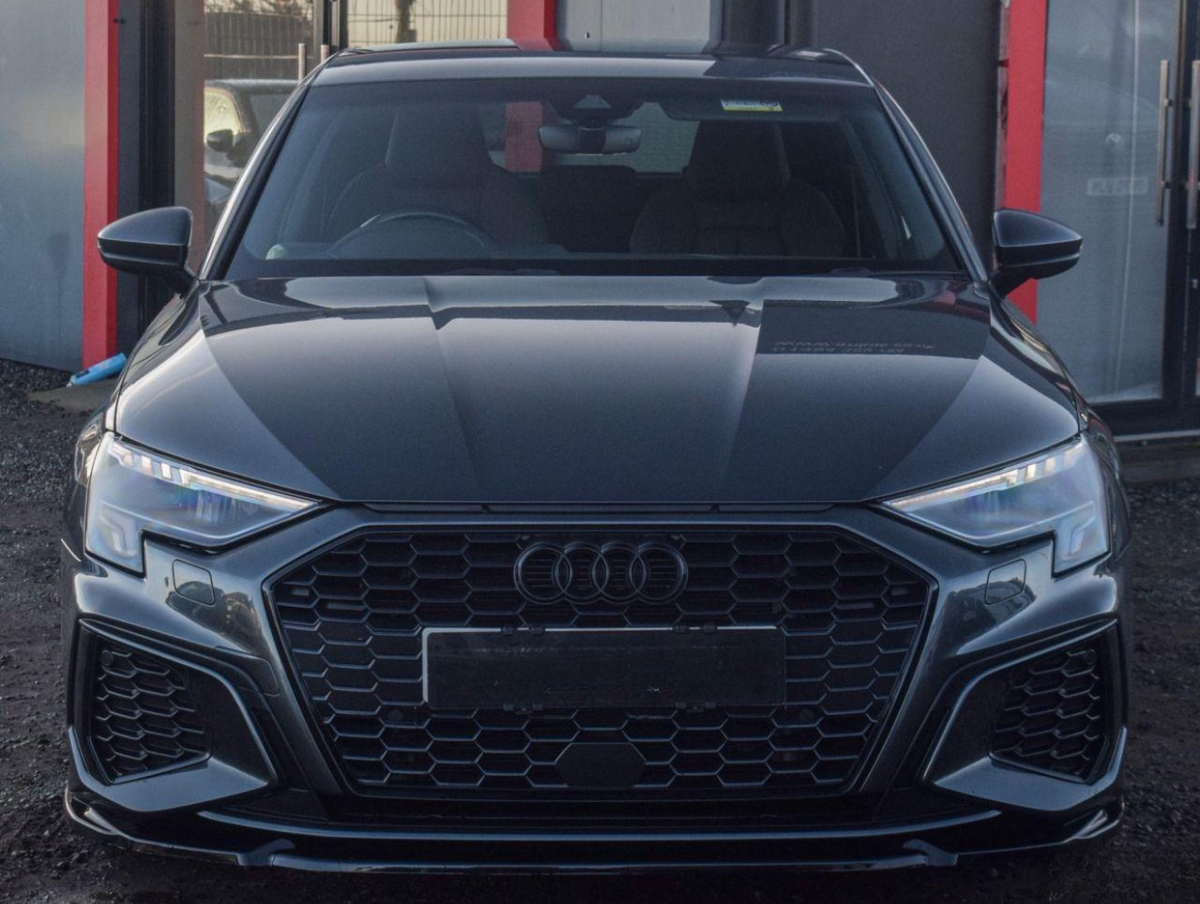 A sleek, black car with a honeycomb grille and sleek headlights is parked on gravel, adjacent to a building with red accents.