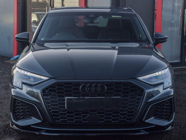 A sleek, black car with a honeycomb grille and sleek headlights is parked on gravel, adjacent to a building with red accents.