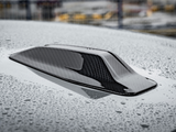 A carbon fiber car roof scoop with raindrops resting on its glossy surface, situated in a wet outdoor area with blurred industrial structures in the background.