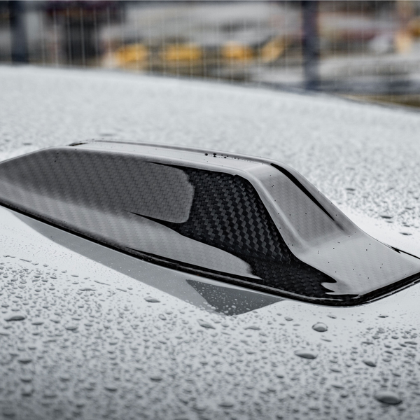A carbon fiber car roof scoop with raindrops resting on its glossy surface, situated in a wet outdoor area with blurred industrial structures in the background.