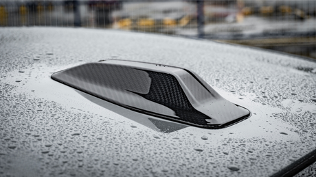 A carbon fiber car roof scoop with raindrops resting on its glossy surface, situated in a wet outdoor area with blurred industrial structures in the background.