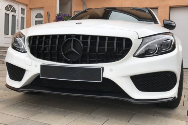 A white Mercedes-Benz car parked on paved ground, displaying a prominent grille with a logo. The background includes a peach-colored building with windows and a door.