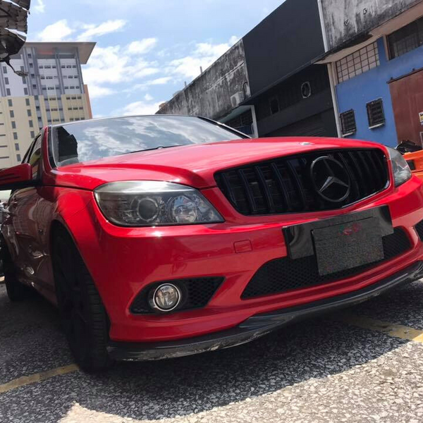 Red car parked on a street with a reflective black grille and black wheels. Surrounded by urban buildings and motorcycles under a partly cloudy sky. The car is positioned diagonally.