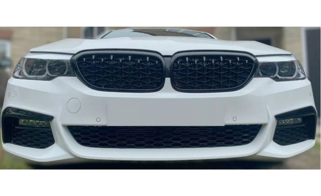 A white car with a prominent front grille and headlights is parked outdoors, facing the viewer. The grille features a black mesh design, framed by a sleek body.