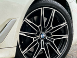 A car's front wheel is stationary, featuring a multi-spoke design with a BMW emblem, against a paved road background. The car is white with visible tire treads.