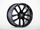 A sleek black alloy wheel is displayed upright, featuring a multi-spoke design with the Tesla logo at its center, set against a plain white background.