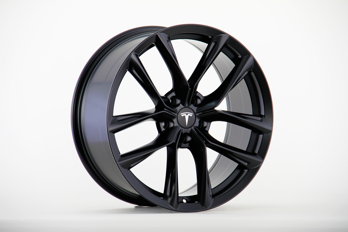 A sleek black alloy wheel is displayed upright, featuring a multi-spoke design with the Tesla logo at its center, set against a plain white background.