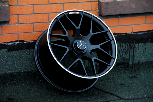 A black alloy wheel with a Mercedes logo rests against an orange brick wall. The wheel features intricate spokes and an "AMG" label, situated on a dark, textured surface.