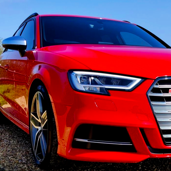 A red sports car with sleek headlights is parked on gravel, under a clear blue sky. The car's distinctive grille and angular design suggest high performance and modern styling.