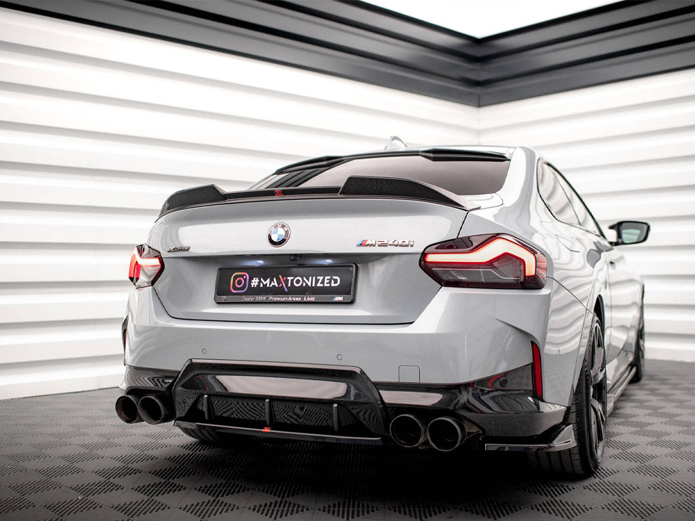 A gray BMW M240i is parked on a checkered floor in a well-lit showroom. The car's rear license plate reads "#MAXTONIZED".