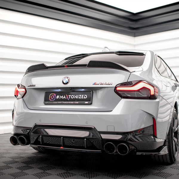 A gray BMW M240i is parked on a checkered floor in a well-lit showroom. The car's rear license plate reads "#MAXTONIZED".