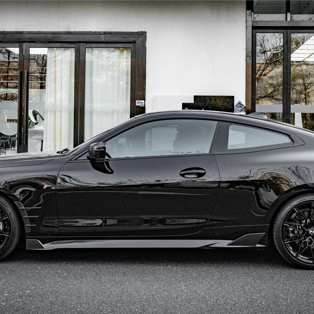 A sleek black sports coupe is parked in front of a modern building with large glass doors and light curtains, reflecting the surrounding trees and architecture.