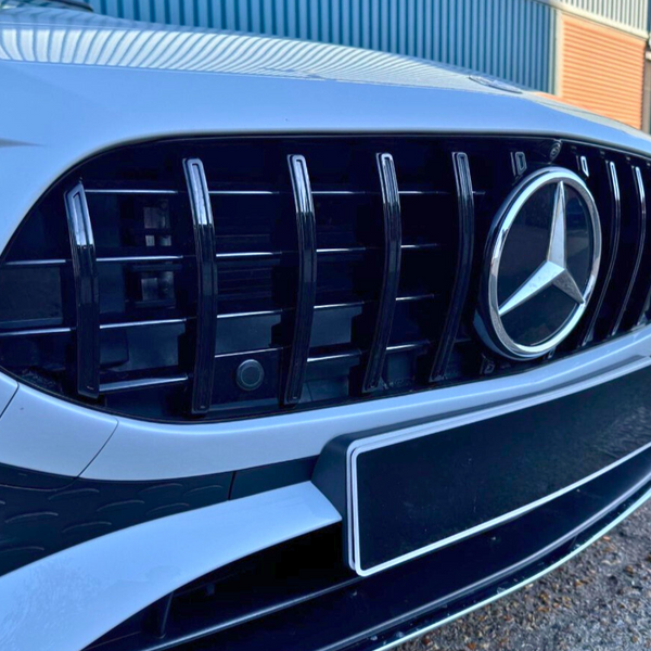 A white car features a prominent black grille with a silver emblem, parked on asphalt near blue and orange industrial buildings.