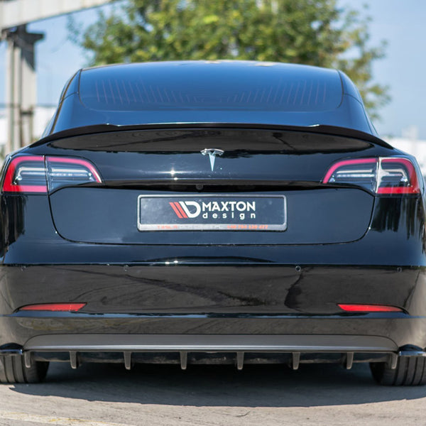 A black car, viewed from the rear, displays a license plate with "MAXTON design" text. It is parked on a concrete surface next to a fence and greenery under a clear sky.