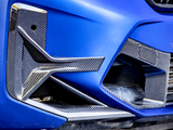 A blue car's front bumper with carbon fiber detailing features aerodynamic fins and a small vent, surrounded by a smooth, glossy finish and adjacent grille.