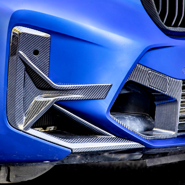 A blue car's front bumper with carbon fiber detailing features aerodynamic fins and a small vent, surrounded by a smooth, glossy finish and adjacent grille.
