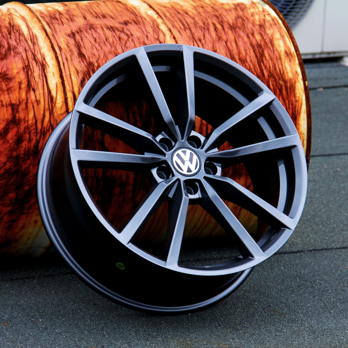 A black Volkswagen alloy wheel is leaning against a large, wooden barrel with a textured orange surface, placed on a gray concrete floor.