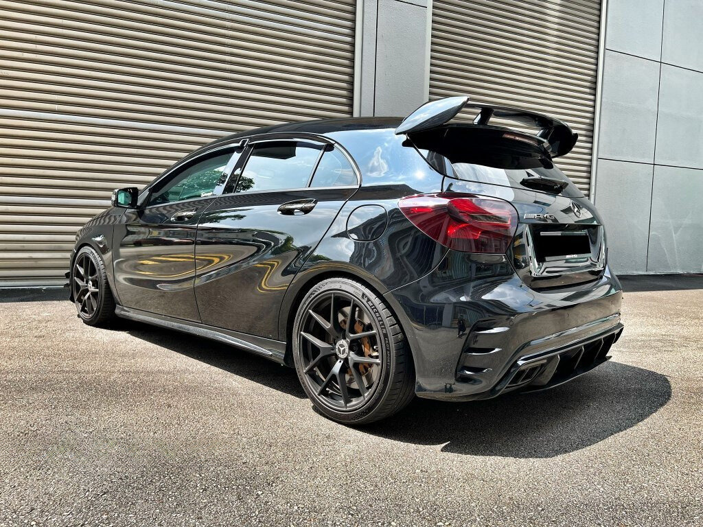 A sleek, black hatchback car, parked on a concrete surface, features sporty wheels and a prominent rear spoiler. It's positioned near a large industrial-style metal garage door.
