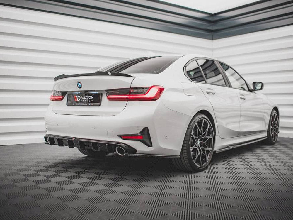 A white BMW sedan featuring sporty black wheels is parked, highlighting its dual exhaust and sharp rear design. The setting is a modern garage with textured walls and flooring. The text on the license plate reads "MAXTON DESIGN."